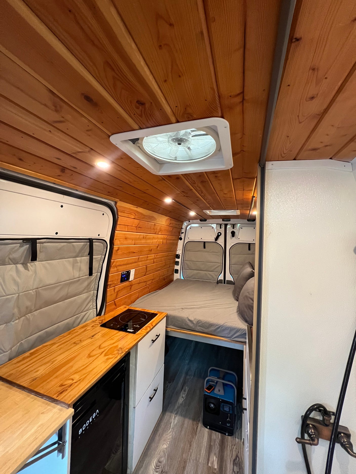 Camper van RV rental interior: wooden ceiling, cozy bed, kitchen with induction cooktop, fridge, and portable power station.