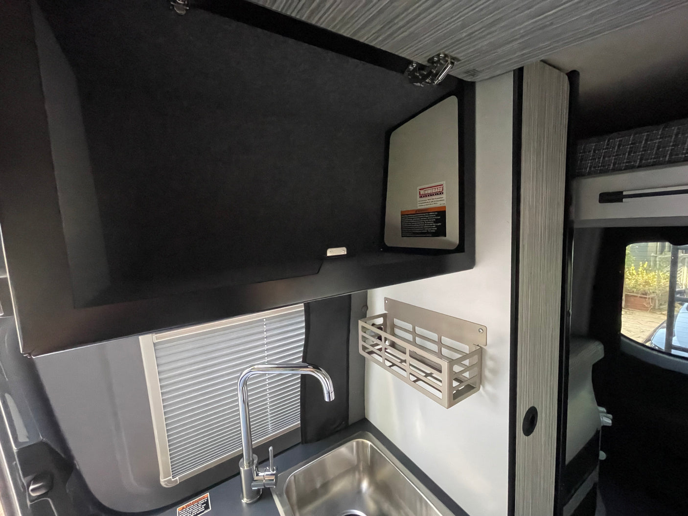 View of a modern RV rental kitchenette with a stainless steel sink, sleek chrome faucet, overhead cabinet, and wall-mounted shelf.