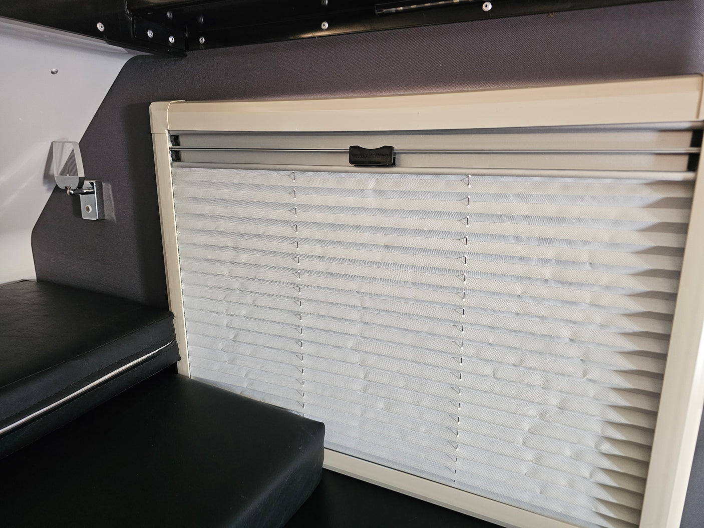 RV rental interior: closed light gray pleated blinds on a window, beige frame. Black cushions below in a compact camper area.