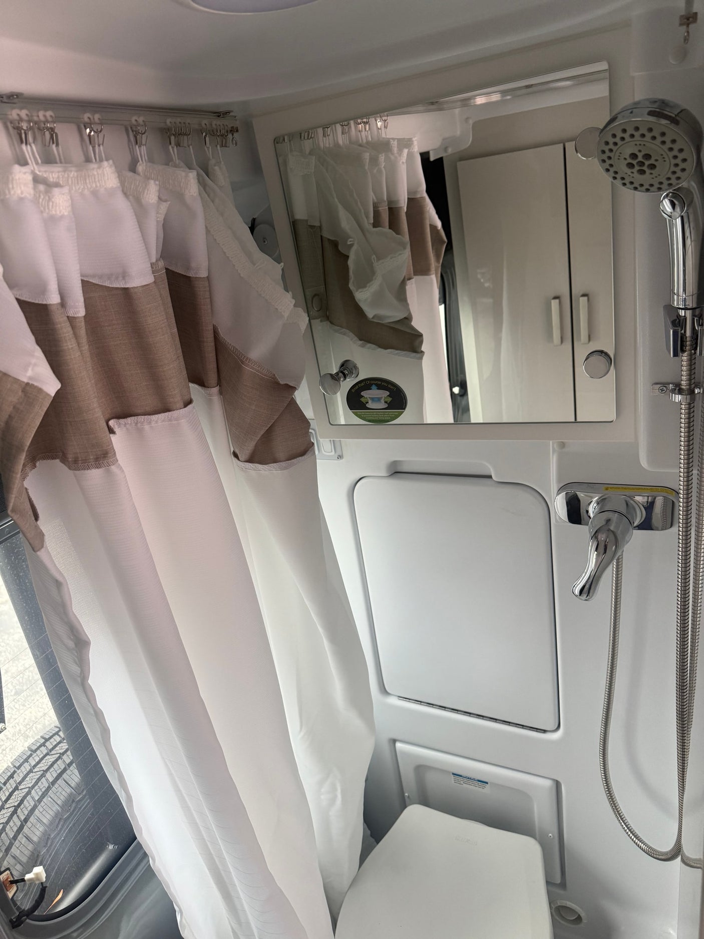 Compact RV rental bathroom with shower, toilet, mirror, and a white/brown curtain by a window showing a tire.