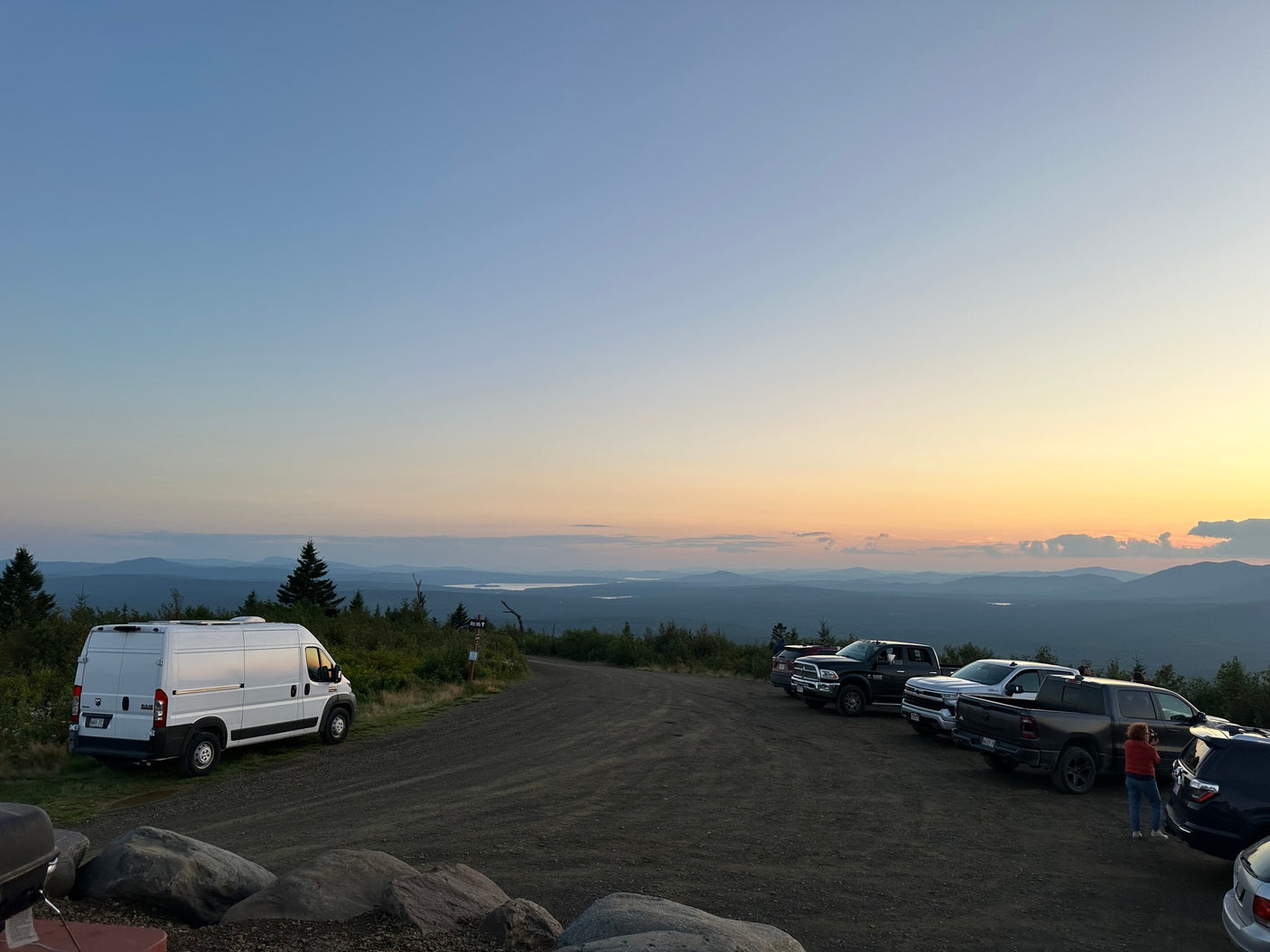 Ram ProMaster RV rental parked at a mountain overlook with a sunset sky over distant lakes and mountains.|A white RV rental camper van is parked on a scenic mountain road under a bright blue sky