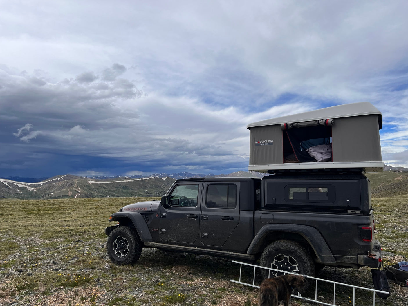 Dark grey Jeep Gladiator with a rooftop tent, an RV rental, overlooking snowy mountains under dramatic cloudy skies.