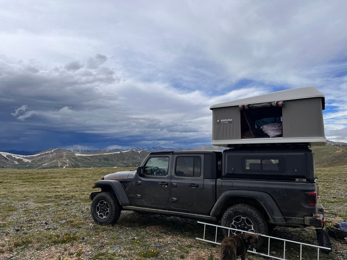 Dark grey Jeep Gladiator with a rooftop tent, an RV rental, overlooking snowy mountains under dramatic cloudy skies.