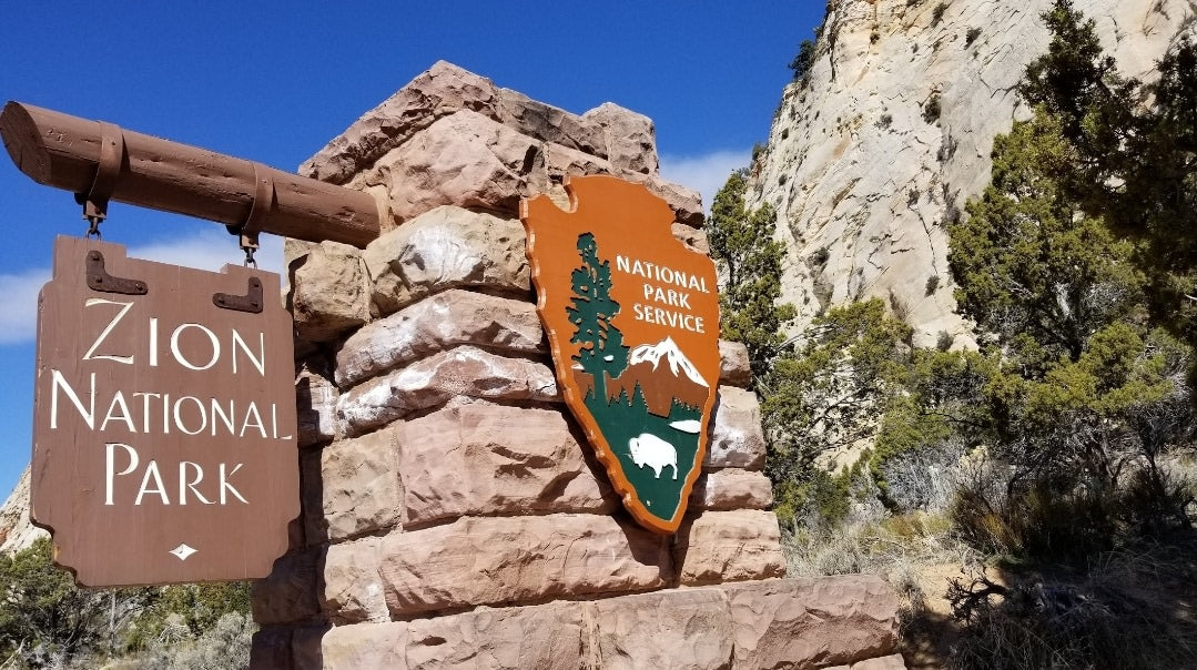 Approaching Zion National Park via RV rental. Iconic entrance sign nestled among stunning cliffs and green foliage awaits your adventure.