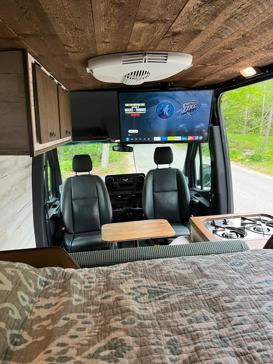 Cozy RV rental interior with a ceiling-mounted TV displaying an NBA game, compact kitchen, and comfortable bed. Perfect for road trips.