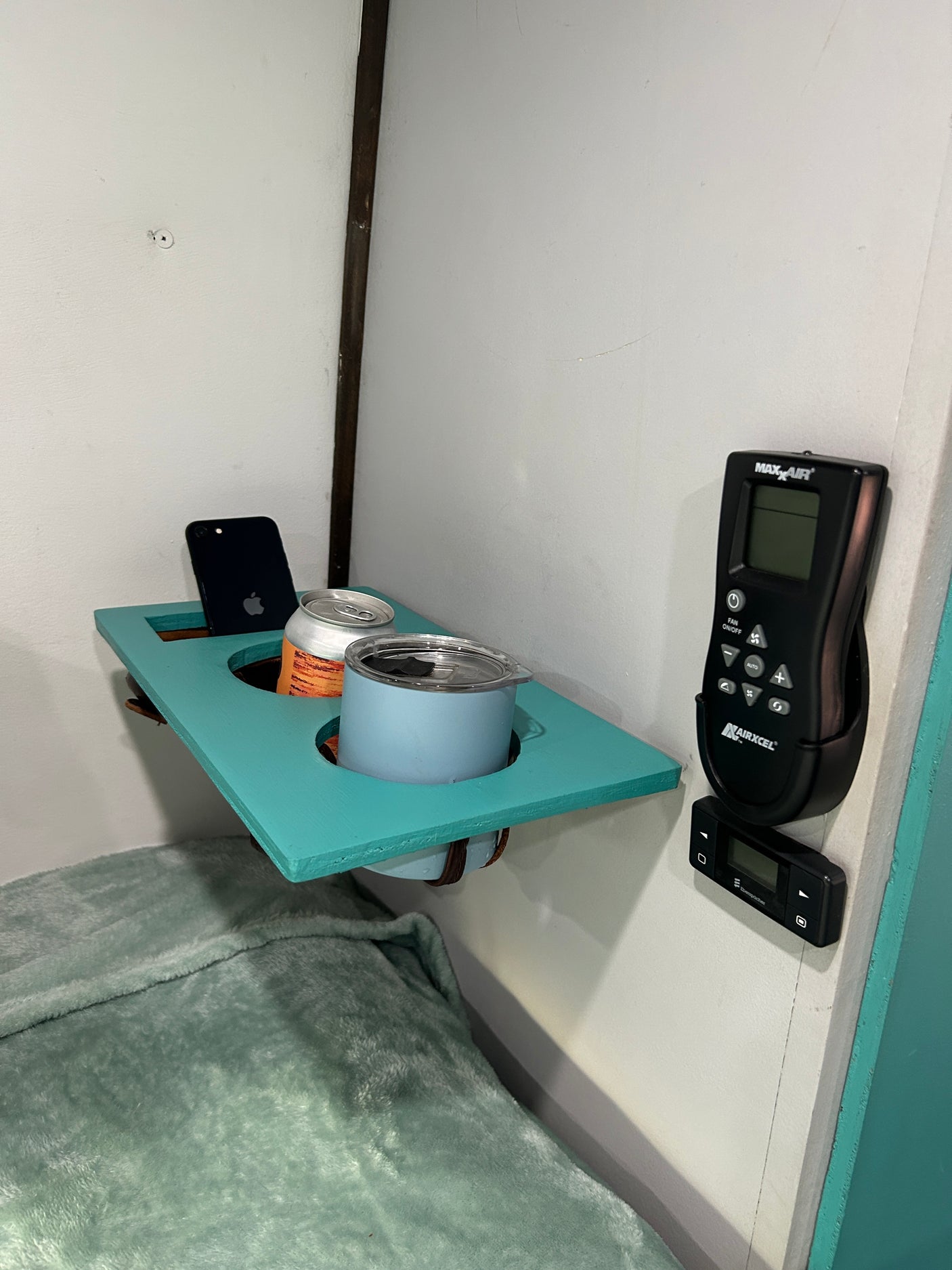 Cozy RV rental bedroom with a teal bedside shelf holding a phone, canned drink, and mug, alongside climate controls.