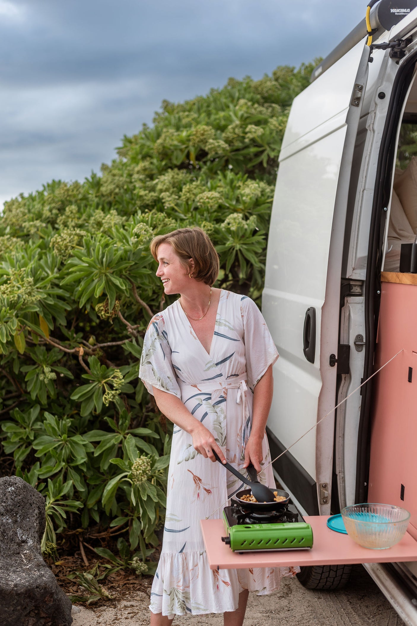 Enjoying an RV rental: woman cooking outdoors with a portable stove next to a white campervan, surrounded by green foliage.