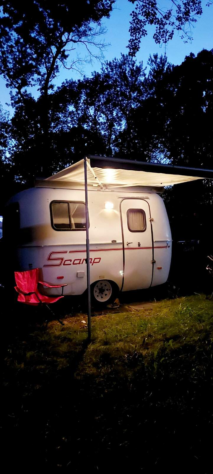 Cozy Scamp RV rental at night, illuminated under an awning with a red chair, set for camping amidst dark trees.