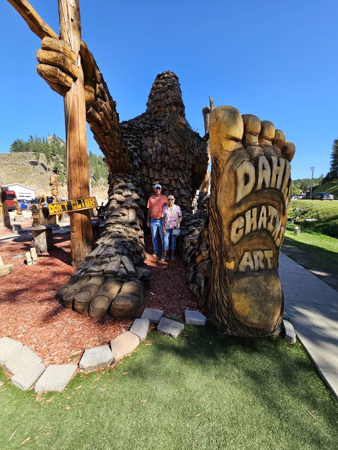 A couple poses with a huge Bigfoot wood carving, featuring Dahl Chat Art. Ideal stop for your RV rental journey!