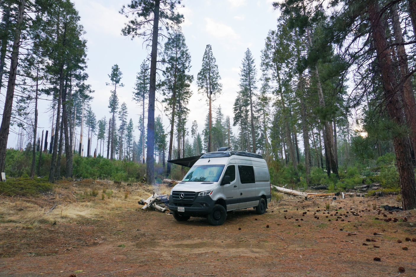 Enjoy nature's beauty with an RV rental. Silver camper van parked in a forest clearing next to a smoky campfire.