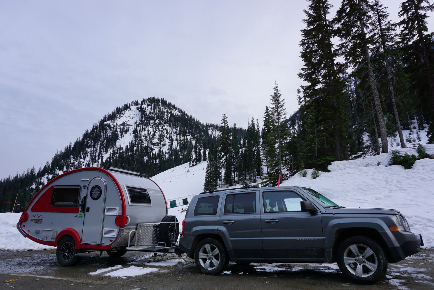 RV rental adventure: Teardrop trailer towed by Jeep in snowy mountains, surrounded by evergreen trees.