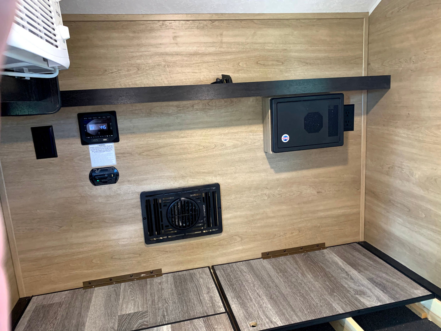 Inside an RV rental: wall-mounted AC, digital control panel, electrical components, and hinged bed base with wood-look flooring.