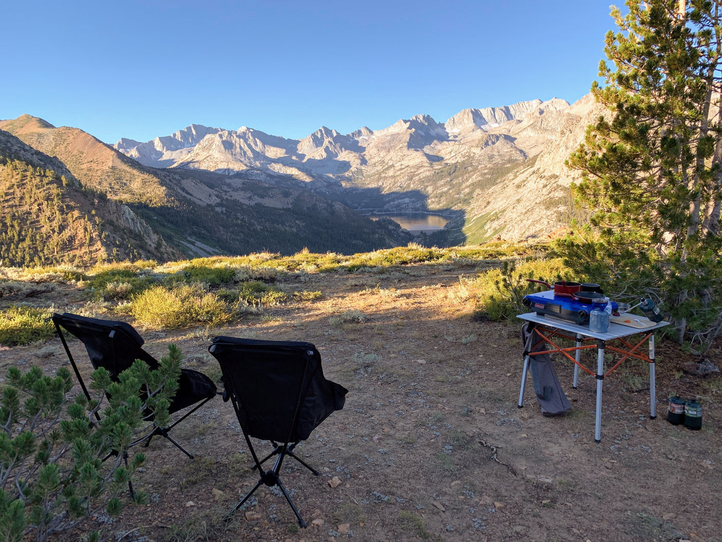 Peaceful RV rental campsite with chairs, stove, table, overlooking a stunning mountain and lake view.