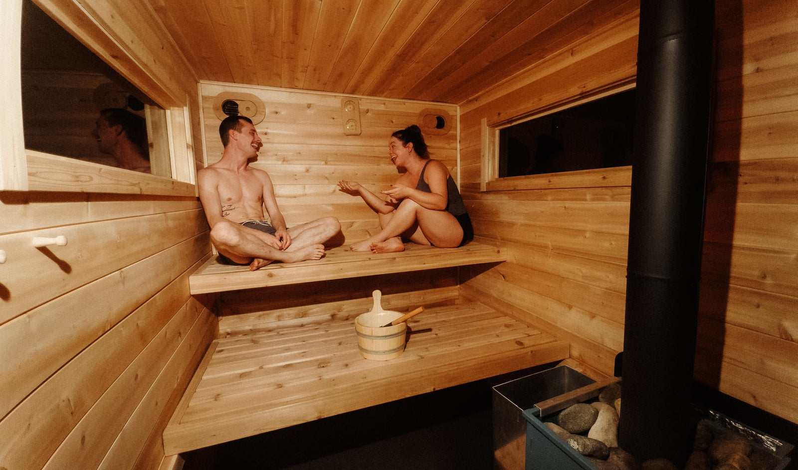 RV rental relaxation: Two people chat and laugh in a cozy wooden sauna with a heater and bucket, perfect for unwinding.