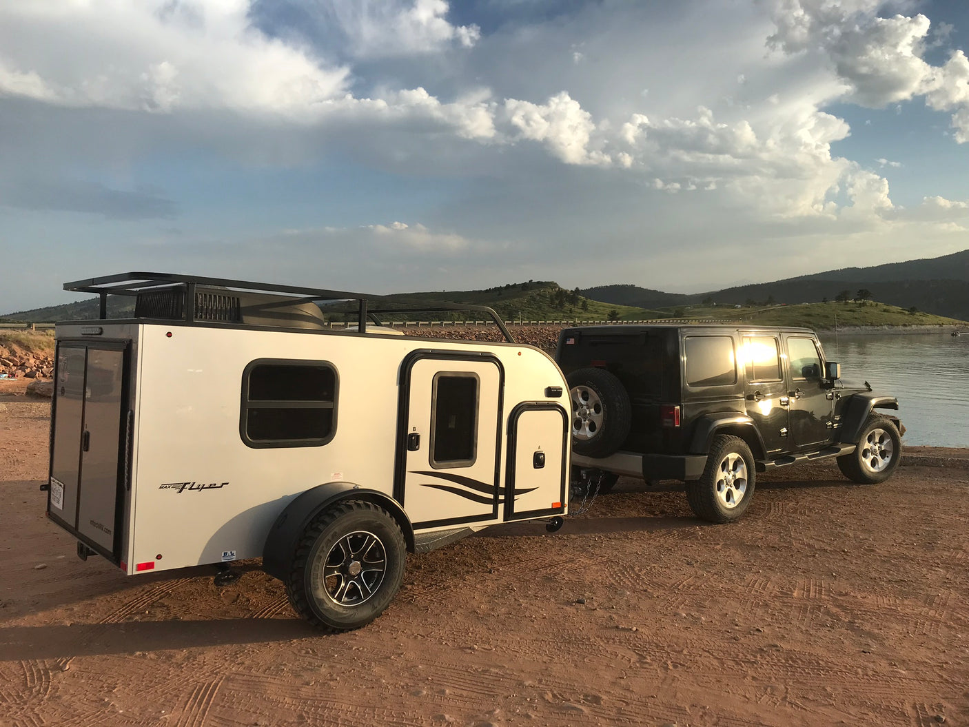 Black Jeep Wrangler towing a white inTech Max Flyer RV, parked lakeside. An ideal RV rental for off-road camping adventures.