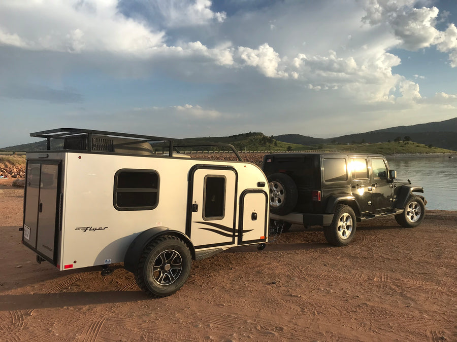 Black Jeep Wrangler towing a white inTech Max Flyer RV, parked lakeside. An ideal RV rental for off-road camping adventures.