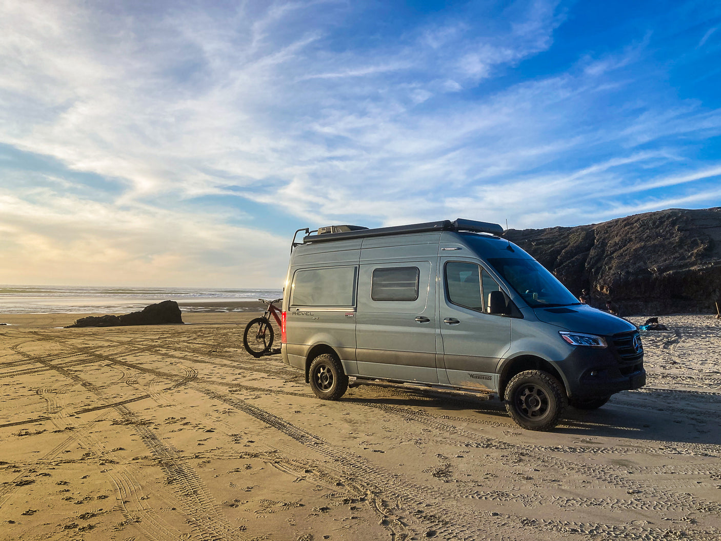 Experience van life! This Winnebago Revel RV, perfect for an RV rental, is parked on a golden sandy beach at sunset with a bike.