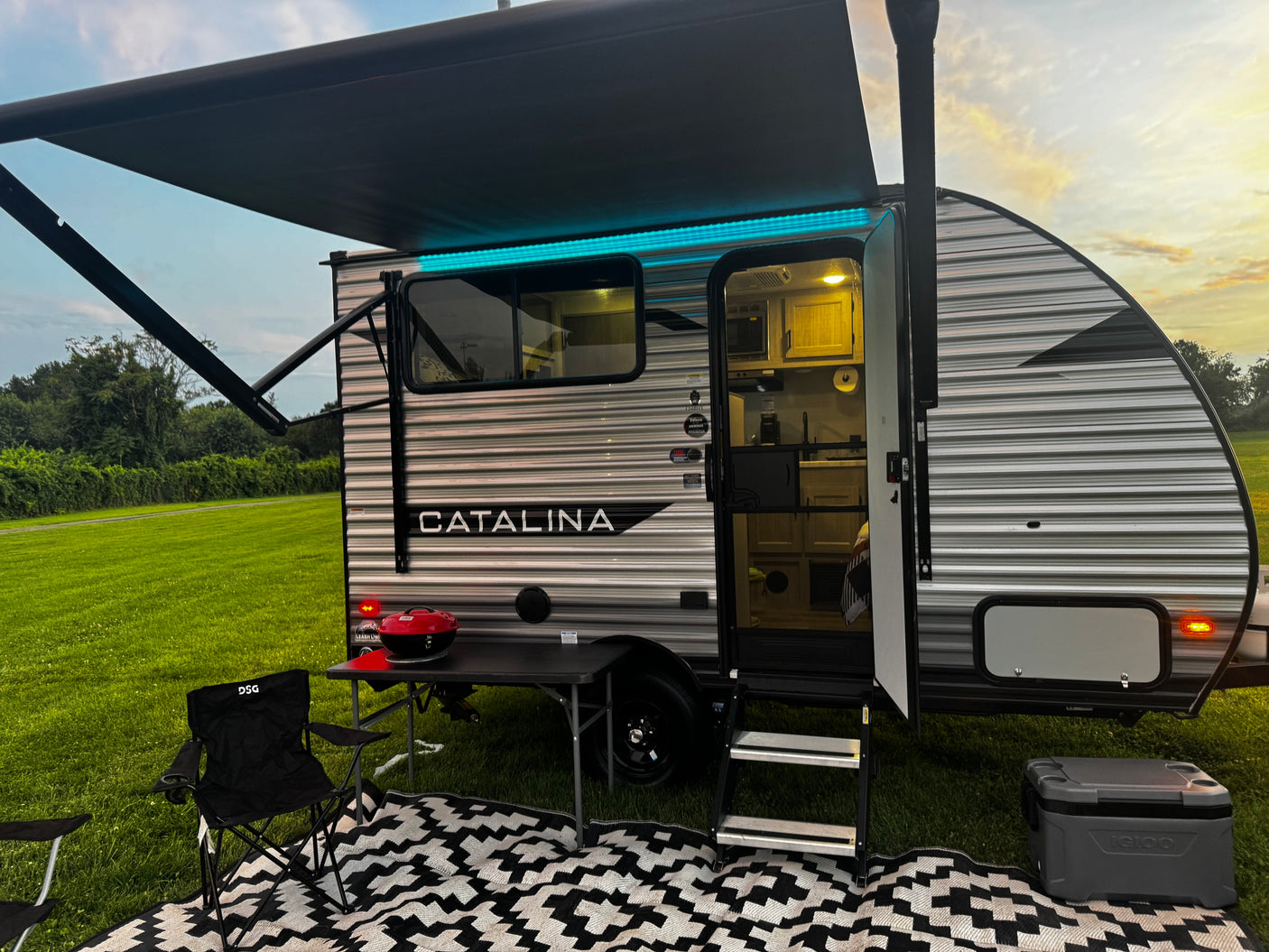 An RV rental travel trailer is set up with an illuminated awning, outdoor mat, grill, and cooler for a relaxing evening.