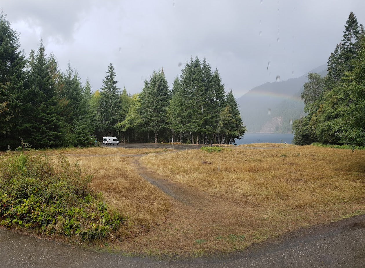 An RV rental parked in a dry grass field surrounded by tall evergreen trees, with a lake and rainbow in distance.