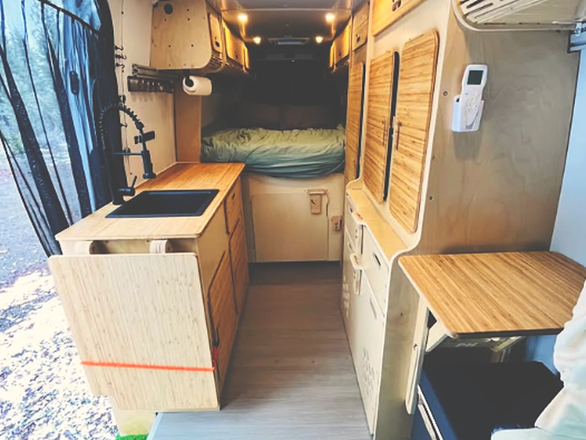 screened interior for winter travel.|View inside an RV rental camper van. Two front seats have vibrant mountain scene covers. Dashboard & outside forest visible.|White campervan (RV rental) with an open side door — photo 8