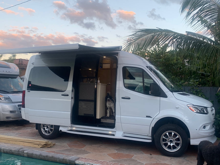 parked on concrete. Sunlight glints off its hood near a building and trees.|White Sprinter RV rental on a patio with two fluffy dogs. A person is seen inside the comfortable camper van.|White Mercedes Sprinter RV rental and a happy black dog on a tropical patio