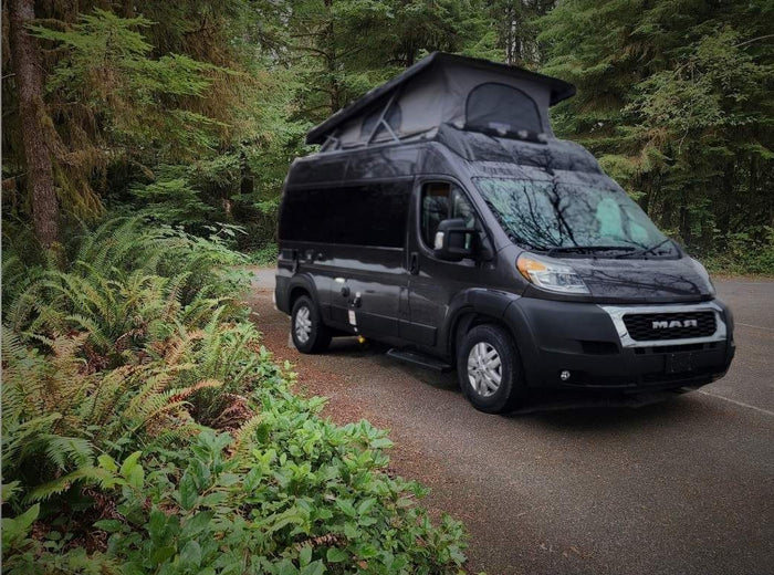Dark grey camper van with pop-top extended, parked by lush forest. Your ideal rv rental for nature adventures.