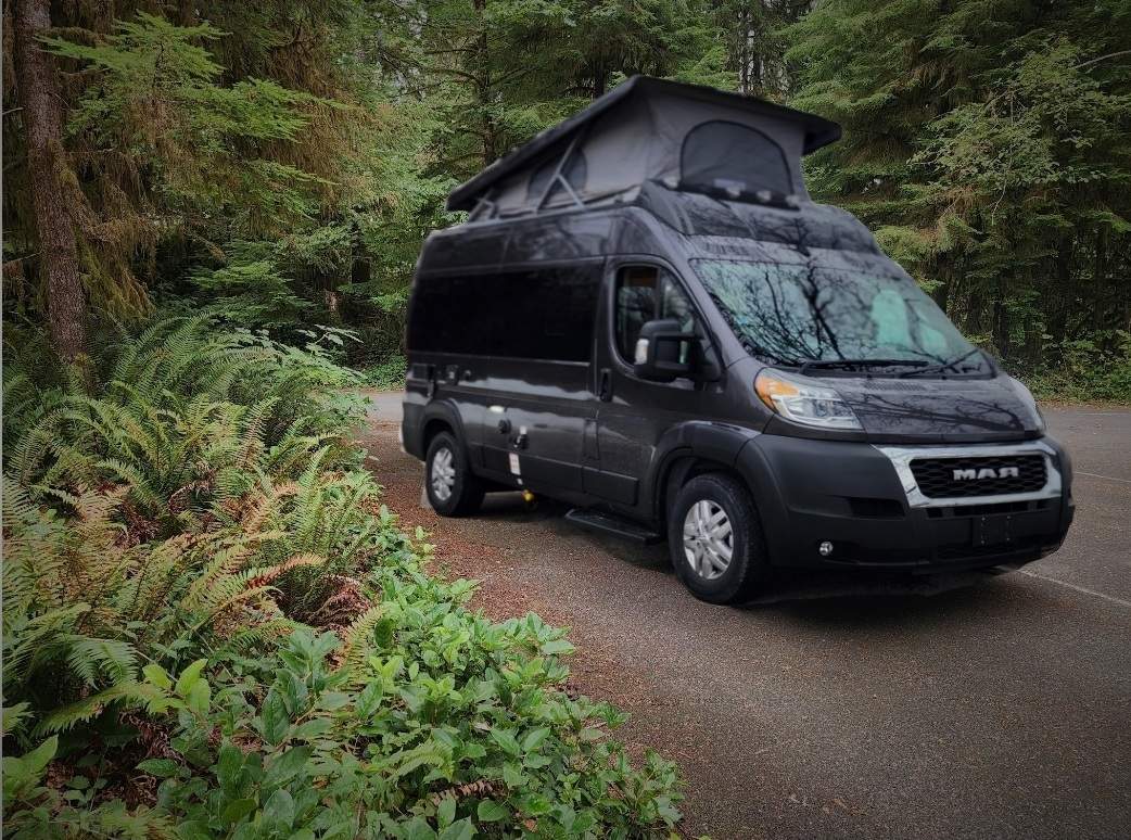 Dark grey camper van with pop-top extended, parked by lush forest. Your ideal rv rental for nature adventures.
