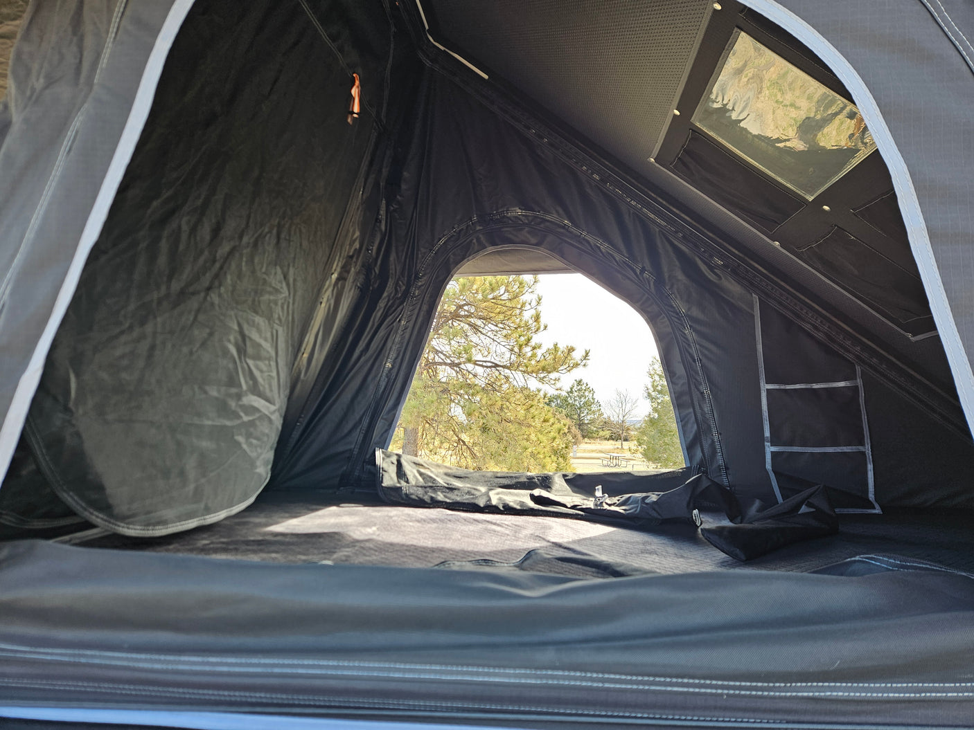 View from inside a dark, spacious rooftop tent, looking out at trees and a picnic area, great for an RV rental adventure.