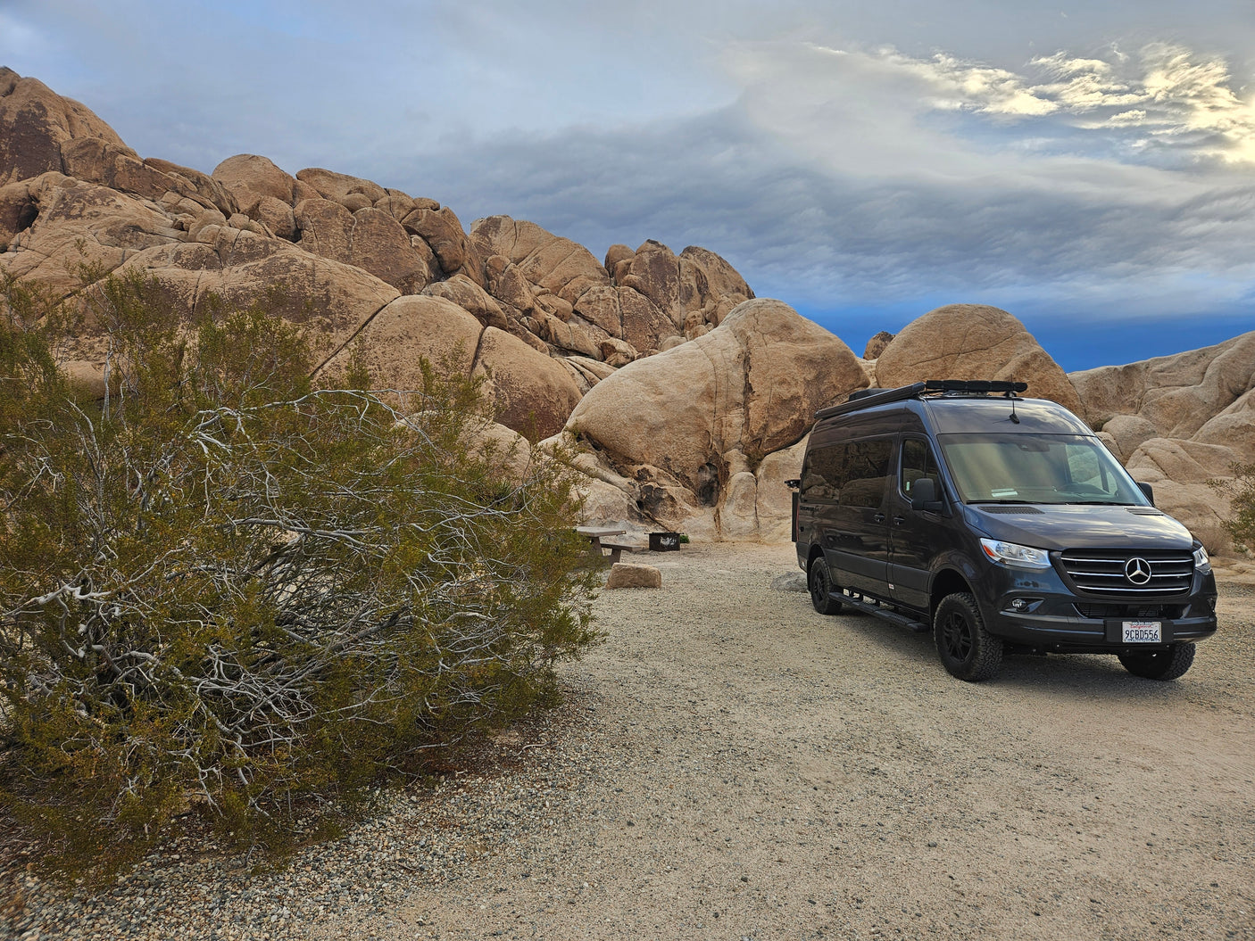 An RV rental, a dark gray camper van, parked at a desert campsite next to large rock formations and a picnic table.