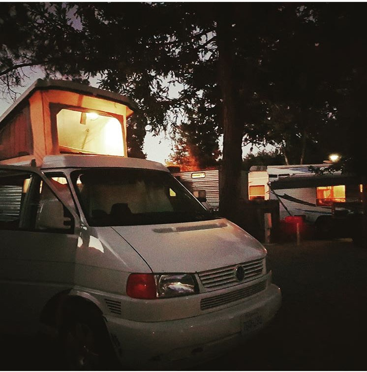A white VW pop-top camper van with an illuminated top, parked at night in an RV park. Perfect for an RV rental adventure.