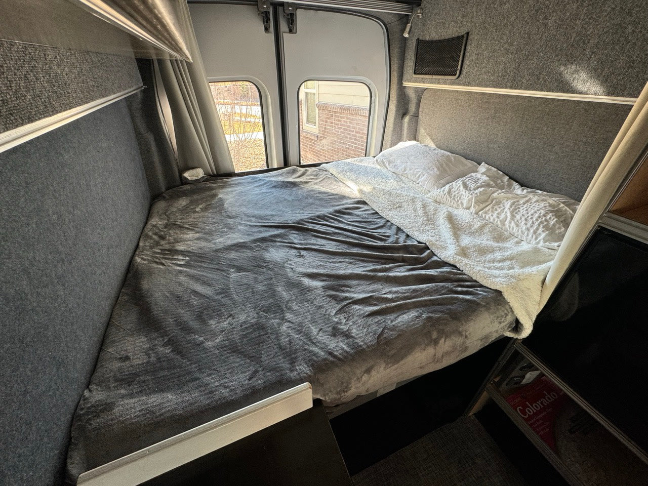 RV rental: comfortable bed in a camper van interior with grey and white bedding. Rear windows show an outside view.