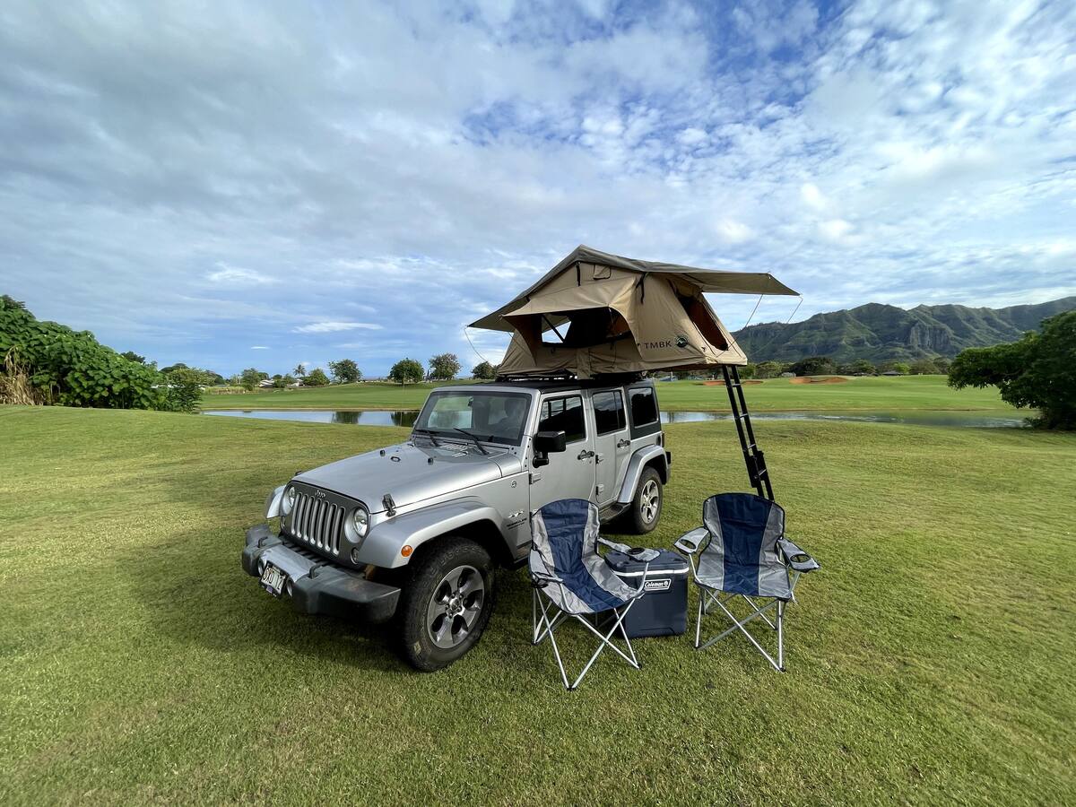 mountains in Hawaii background. Ideal for an RV rental adventure.|Adventure-ready silver Jeep with TMBK rooftop tent & camp chairs on grassy field. Explore with our RV rental!|An ideal beach camp setup: two chairs