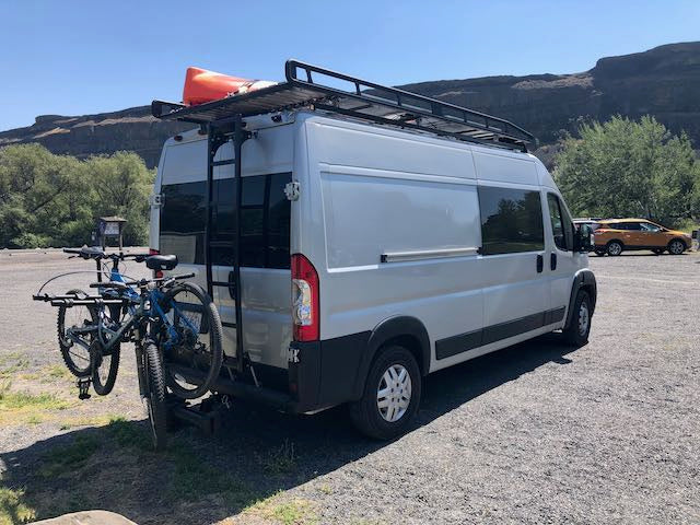 White RV rental van with bikes and kayak on its roof rack, parked in a sunny mountain landscape, ready for outdoor adventure.