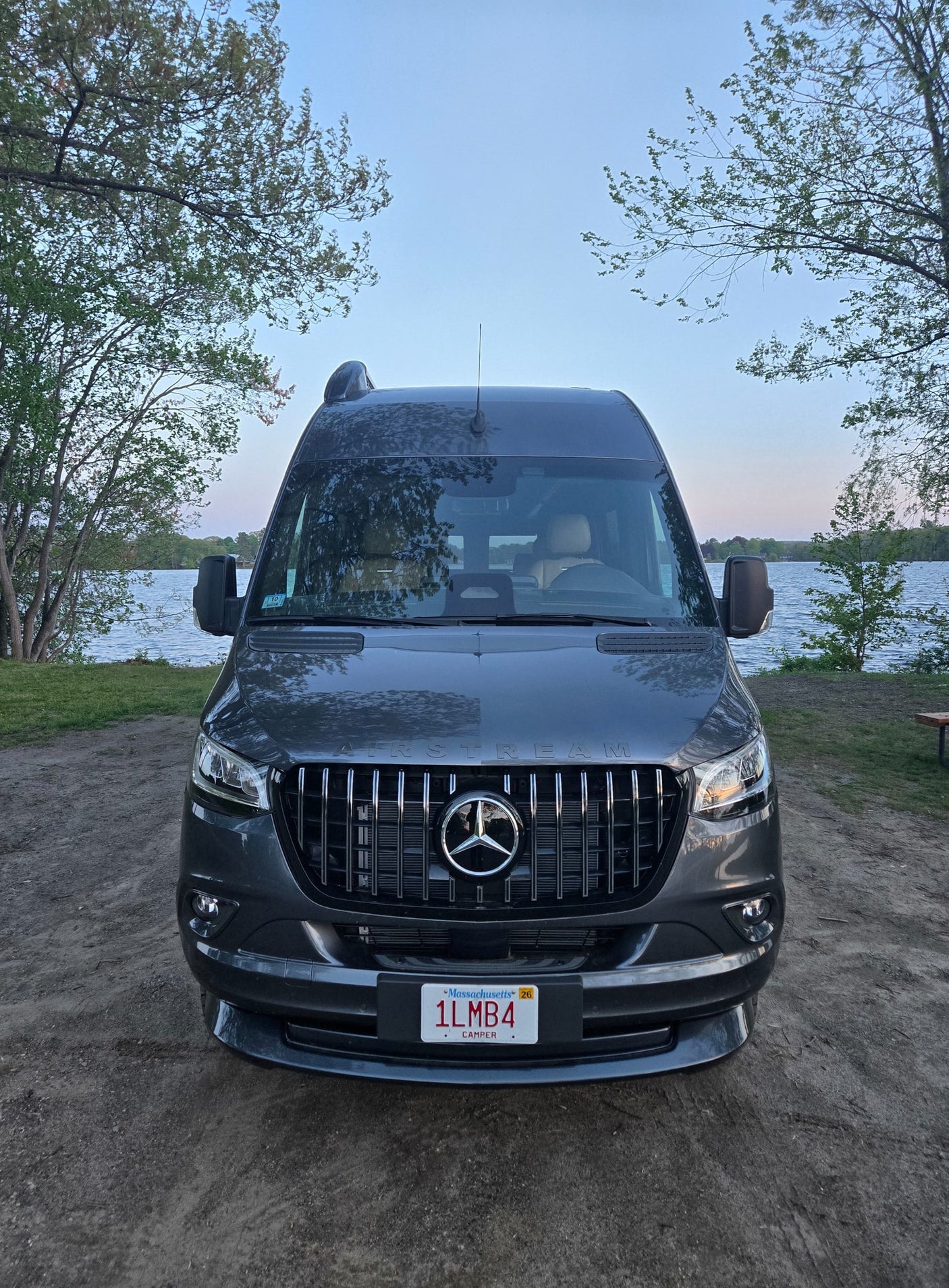 Dark gray Airstream rv rental camper van parked by a lake under a clear sky, surrounded by trees. Ideal for outdoor adventures.