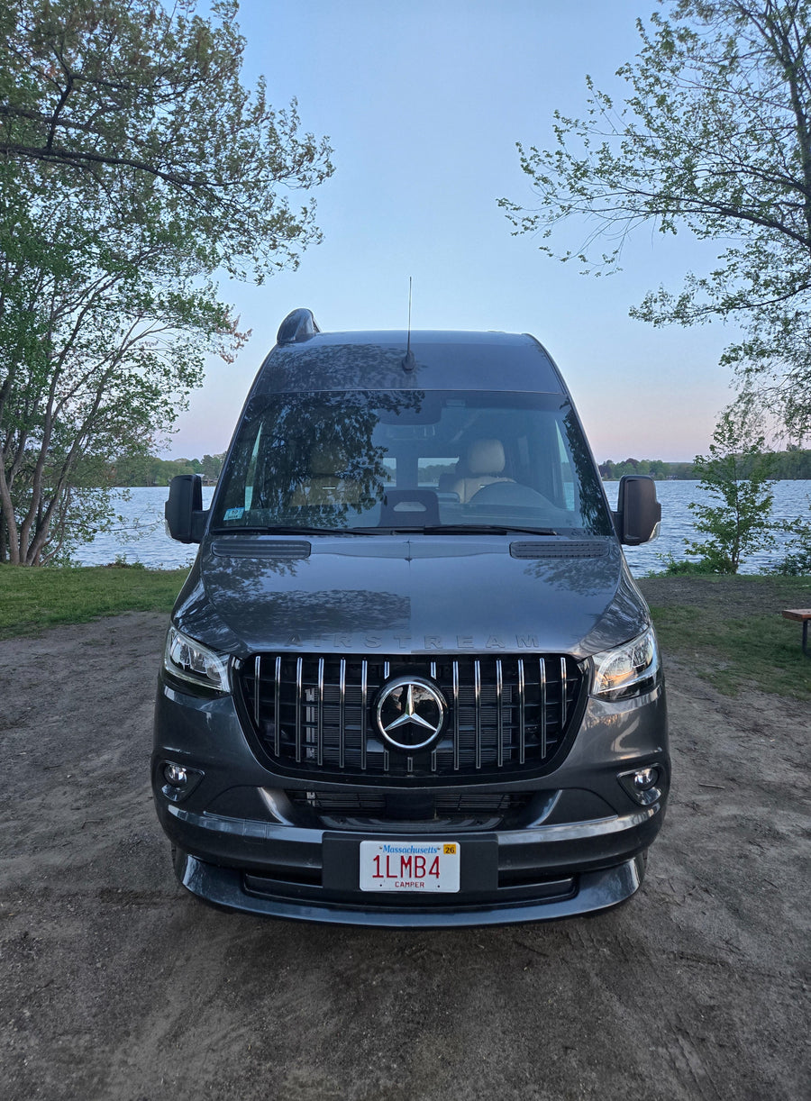 Dark gray Airstream rv rental camper van parked by a lake under a clear sky, surrounded by trees. Ideal for outdoor adventures.