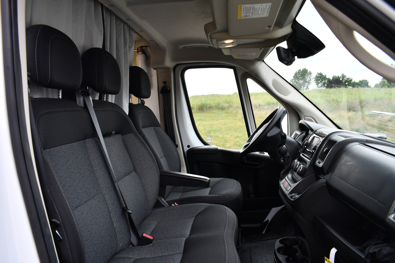 Inside an RV rental van: front seats, dashboard, and cabin, with curtains & fire extinguisher, overlooking a field.