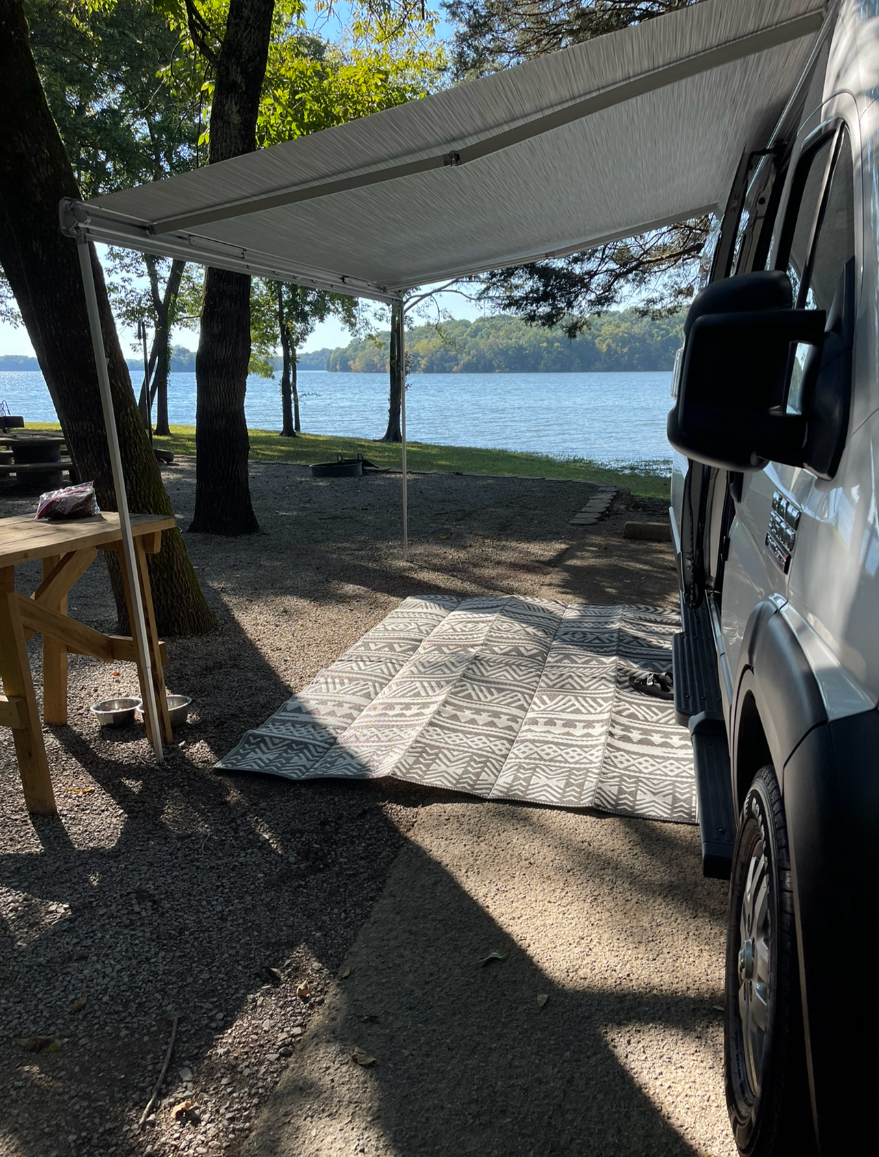 Relaxing RV rental setup by the lake with an extended awning, mat, and picnic table at a scenic campsite.