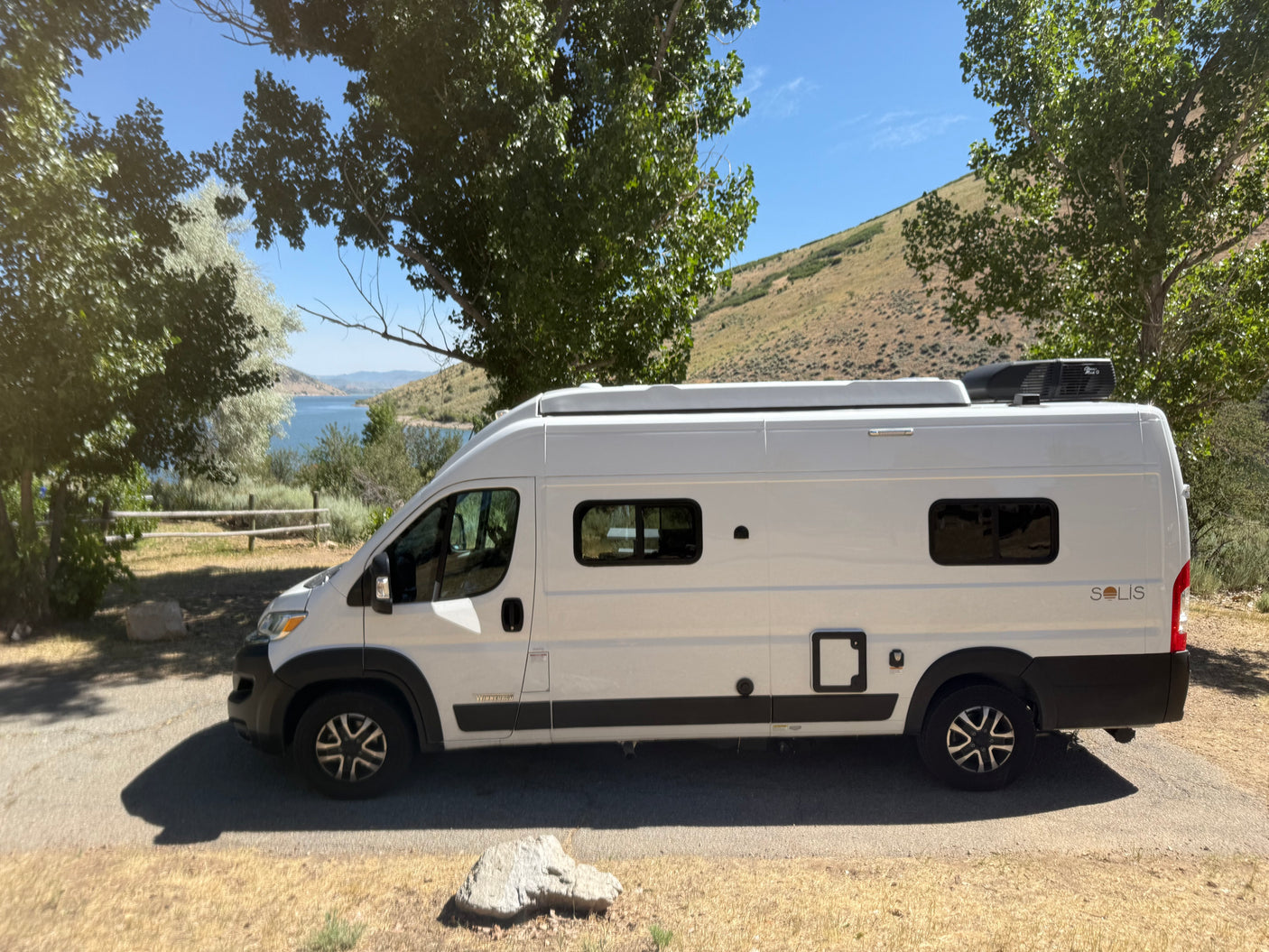 A white Winnebago Solis camper van, an ideal RV rental, parked by a scenic lake and hills. Perfect for travel adventures.