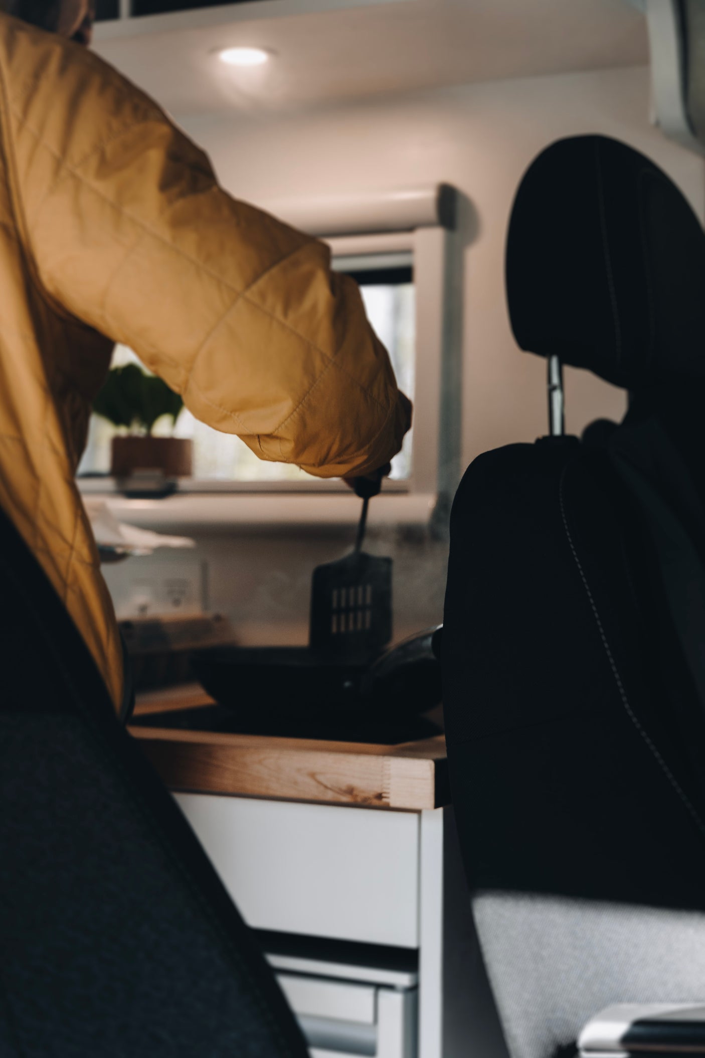 Person cooking in an RV rental kitchen, stirring food in a steaming pan. A vehicle seat is in the foreground.
