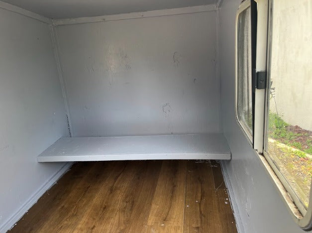 RV rental interior: a clean, grey-painted compartment with a fixed shelf, laminate floor, and natural light from a window.