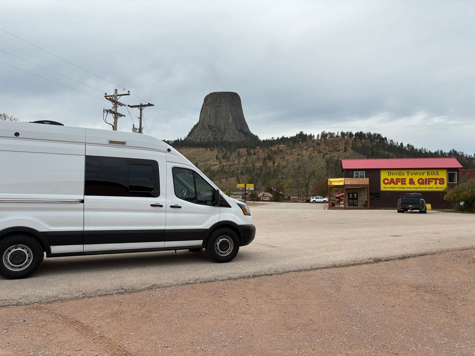 White RV rental van parked at Devils Tower KOA Cafe & Gifts with Devils Tower in the background under an overcast sky.