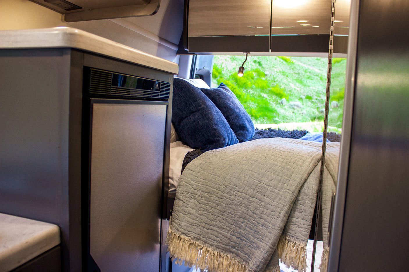 Compact RV rental interior features bed with blue pillows, a fridge, and scenic green views outside.