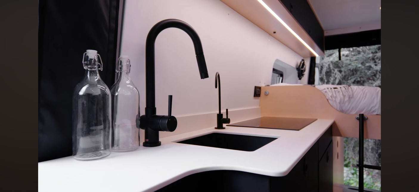 Sleek RV rental kitchen area: black sink, faucet, induction cooktop, white counters, and glass bottles. Compact and functional.