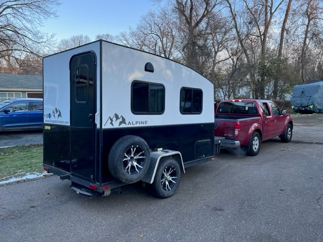 An Alpine RV rental camper, black & white, is hitched to a red pickup truck. This travel trailer is ready for trips.