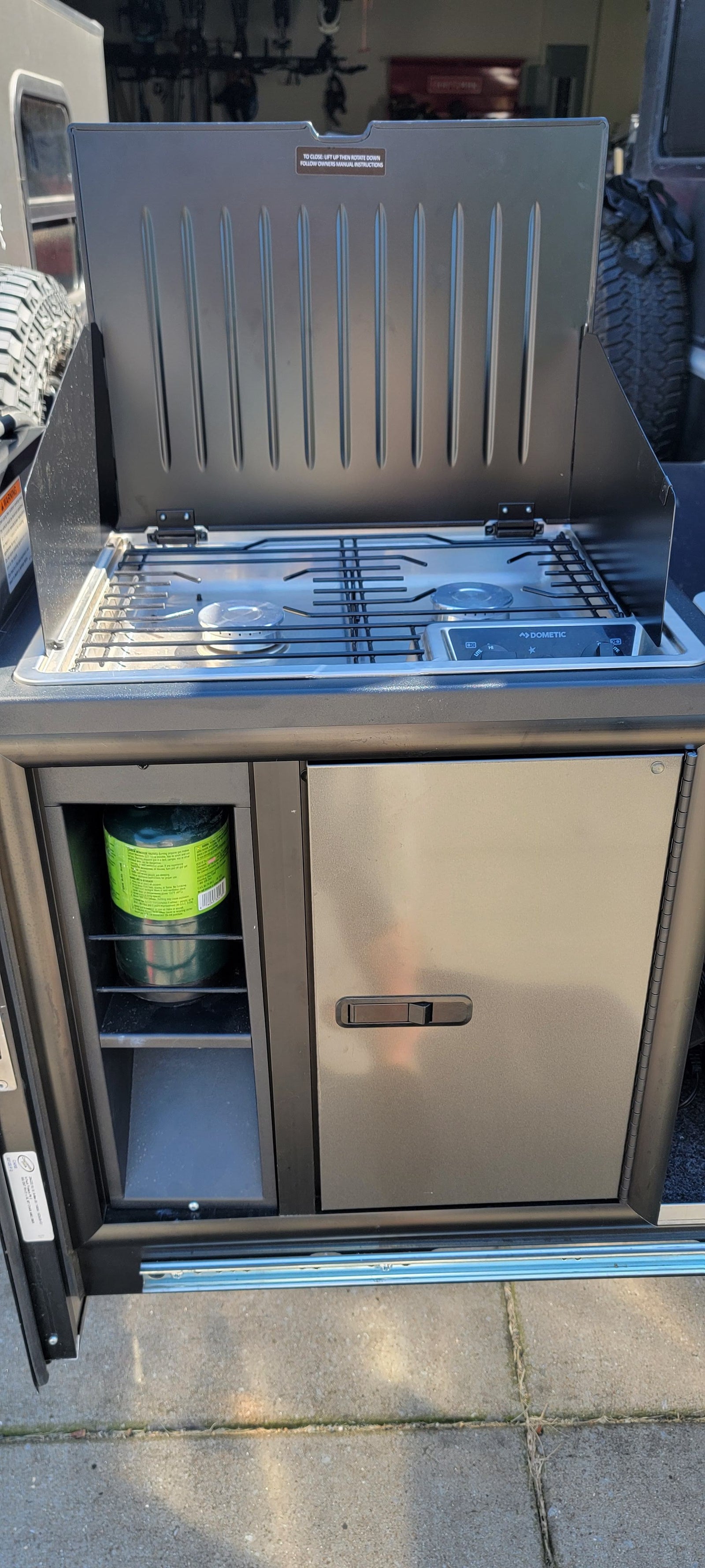 RV rental's slide-out outdoor kitchen with an open Dometic 2-burner gas stove, visible propane tank, and mini-fridge.