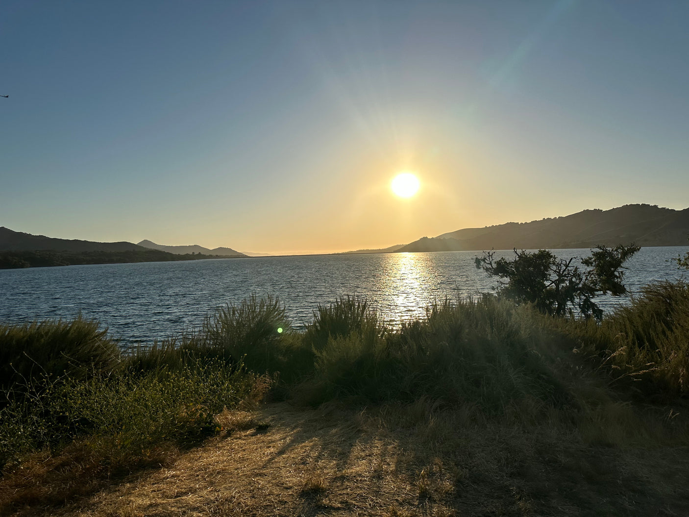 Beautiful sunset over a calm lake, with hills and foreground plants. Perfect scenic stop for your RV rental.
