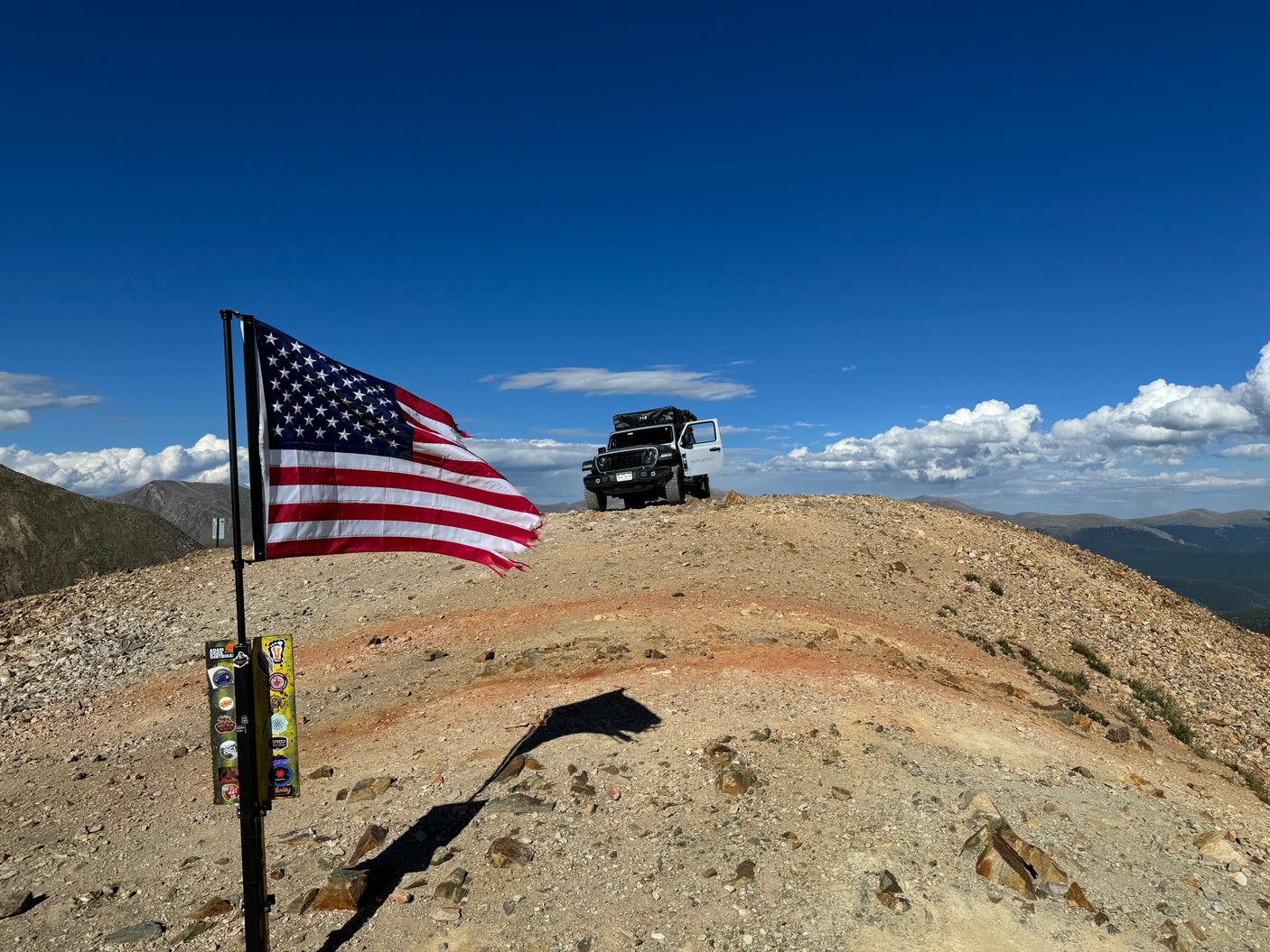 Off-road adventure with an RV rental: a Jeep with a rooftop tent on a rocky mountain peak, American flag in foreground.