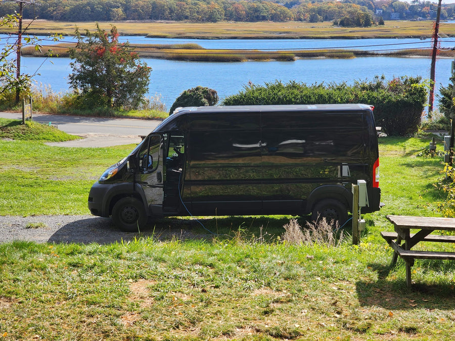 connected to power. A scenic stop for van life camping and outdoor adventure.|Black RV rental van at a damp campsite with picnic tables