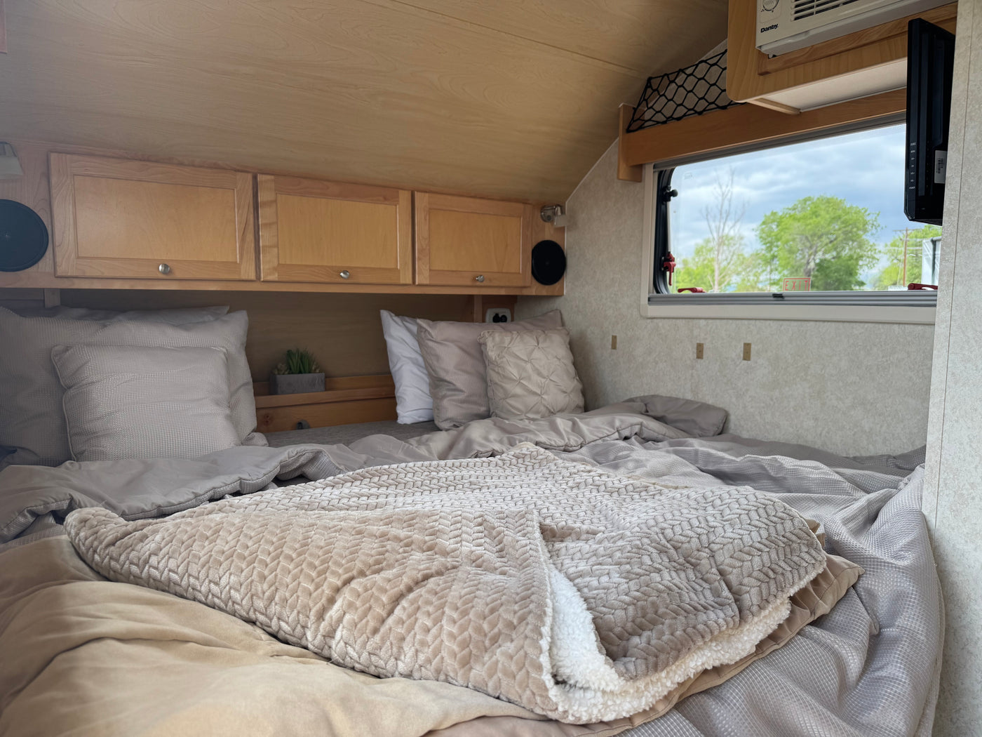 Inviting RV rental interior: a plush bed with neutral pillows/blankets, wood cabinets, and window view for a cozy camping trip. — photo 17