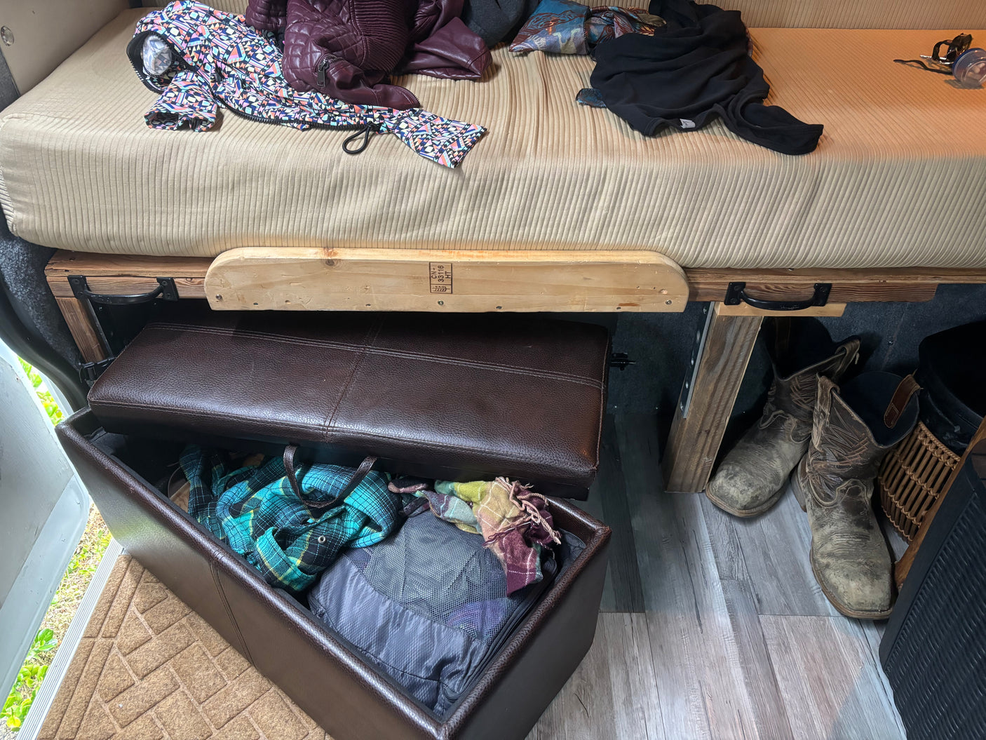 RV rental interior setup: A comfy bed, an open storage ottoman for essentials, and boots on the wood-look floor.
